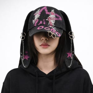 以下が含まれることがあります： ピンク色のグラフィックデザインとピンク色の「DOO」の文字が入った黒いデニムキャップ。キャップには、銀色のチェーンとリングのディテールが付いた、長い耳のようなエクステンションが付いています。ピンクの星も見えます。