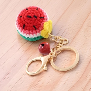 Peut inclure: Un porte-clés fantaisie en forme de pastèque. La pastèque crochetée présente des détails rouges, blancs, verts et noirs. Il comprend un fermoir en métal doré, une perle rouge et un accent de fleur jaune.