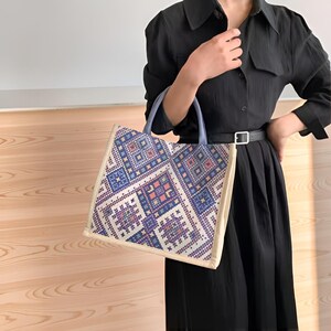 Peut inclure: Un sac fourre-tout avec un motif géométrique dans les tons de bleu, violet et pêche. Le sac a une bordure crème et des poignées bleu clair. Le sac est tenu par une personne portant une robe noire.