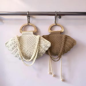 Peut inclure: Deux sacs à main en crochet avec des poignées en bois sont suspendus à une barre métallique. L'un est blanc cassé, l'autre est marron. Les deux ont un bord festonné et une sangle perlée. Les sacs sont exposés contre un mur rose clair.