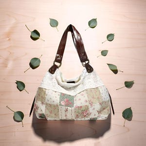 Peut inclure: Un sac à main patchwork crème avec des motifs floraux et une bordure en dentelle. Il est doté de lanières en cuir marron et est posé sur un fond en bois clair avec des feuilles vertes. Le sac présente divers motifs floraux.