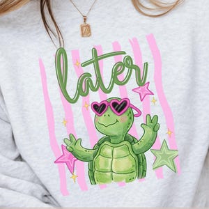 Puede incluir: Sudadera gris claro con una tortuga de dibujos animados con gafas de sol en forma de corazón y la palabra "later" en verde. Rayas verticales rosas y estrellas rodean la tortuga. El diseño incluye el texto "ENGDESIGNPRINTS Original Design" y "PNG FILE INSTANT DOWNLOAD".