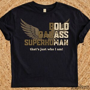 Può includere: Una t-shirt nera a maniche corte con un design grafico raffigurante due ali e il testo BOLD BADASS SUPERHUMAN in un carattere beige dall'aspetto usurato. Sotto la grafica, il testo recita that's just who I am! in un carattere bianco più piccolo.