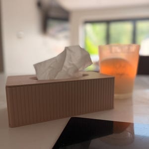 May include: A rectangular, dusty rose-coloured tissue box cover with a vertical ribbed texture. A single white tissue is pulled through the top opening. The cover is positioned next to a glass candle holder with a warm orange glow and a black object.