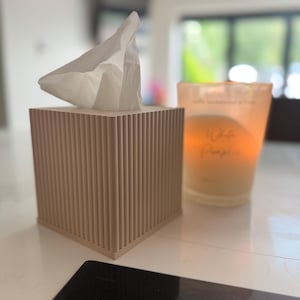 May include: A square, light beige tissue box cover featuring a vertical ribbed texture on all four sides. A single white tissue is pulled through the top opening. Next to the tissue box is a glass candle holder with a warm, glowing orange light.