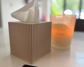 May include: A square, light beige tissue box cover featuring a vertical ribbed texture on all four sides. A single white tissue is pulled through the top opening. Next to the tissue box is a glass candle holder with a warm, glowing orange light.
