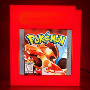 May include: A red, illuminated Nintendo Game Boy cartridge featuring the Pokémon logo and a Charizard illustration. The cartridge has the text "Nintendo GAME BOY" and "THIS SIDE OUT". The game is rated "EVERYONE" by ESRB.