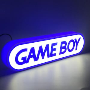 May include: An illuminated sign with the words "GAME BOY" in bold, blue letters. The sign has a white background and a blue border. The sign is lit, and the background is a dark blue. The overall design has a retro feel.
