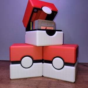 May include: Three red and white storage boxes, designed to look like Poké Balls, are stacked. One is open, displaying game cartridges. The boxes have a white centre, a red top, and a black band. A white circle is on the front.