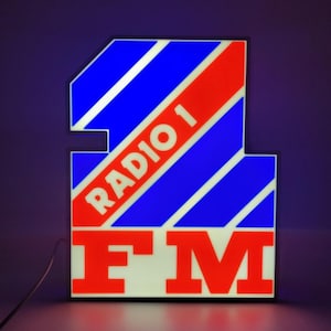 May include: Illuminated sign with the text "RADIO 1 FM". The sign has a blue and red striped design with white accents. The letters are bold and in a sans-serif font. The sign is rectangular with a unique shape.