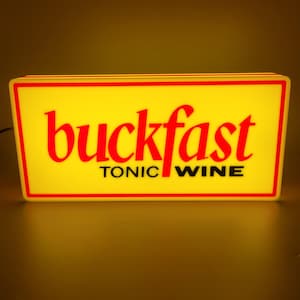 May include: A rectangular illuminated sign with a yellow background and red trim. The sign displays the word "buckfast" in red, stylized lettering above the words "tonic wine" in black.