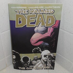 Könnte beinhalten: Ein Comic mit dem Titel "The Walking Dead" wird gezeigt. Das Cover hat einen lila Hintergrund mit dem Titel in Gold und eine grafische Illustration einer Person und Zombies. Der Text "The Calm Before" ist unten sichtbar.