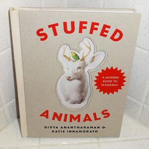 Op de afbeelding: Een boek getiteld "STUFFED ANIMALS" met een illustratie van een hert en een groene vogel. De kaft van het boek is beige met rode tekst en een rode ster met de tekst "A MODERN GUIDE TO TAXIDERMY". De namen van de auteurs staan onder de titel.