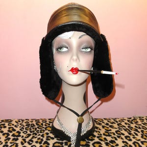 May include: A black leather trapper hat with black faux fur ear flaps and a chin strap featuring a circular gold-tone toggle. The hat is displayed on a mannequin head wearing a silver-tone rhinestone necklace and a long black cigarette holder.