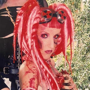 May include: A person with red hair and makeup, wearing goggles and a black top, with a prosthetic rib cage and fake blood. The image has a horror theme.