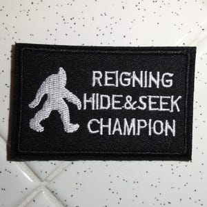 Puede incluir: Parche rectangular negro con texto bordado blanco y silueta de Pie Grande. El texto dice "REIGNING HIDE&SEEK CHAMPION". El parche está sobre una superficie moteada blanca y gris.