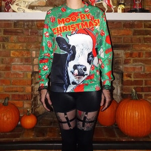 May include: Green Christmas sweater with a cow wearing a Santa hat and the text "Moo-ry Christmas!". The sweater has a festive design with red and white accents. The person is wearing black leggings and fishnet stockings.