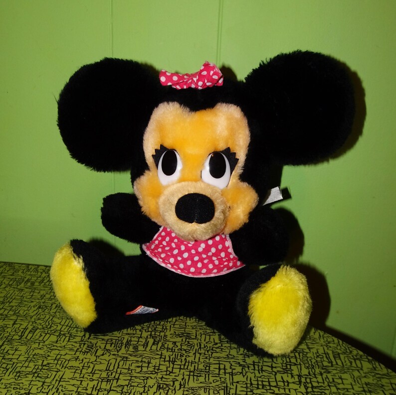 May include: A plush Minnie Mouse doll with black fur, large round ears, and a yellow face. She wears a red and white polka dot bow and matching collar. Her feet are yellow. The doll is sitting on a green patterned surface.