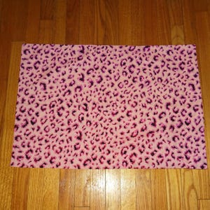 May include: A rectangular pink fabric with a leopard print pattern. The pattern features dark purple and black spots scattered across the pink background. The fabric is likely used for crafting or home decor.