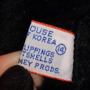 May include: A white label with blue text and an orange border is held against a black background. The label reads "HOUSE OF KOREA" and "LIPPINGS TENELLS KEY PRODS."