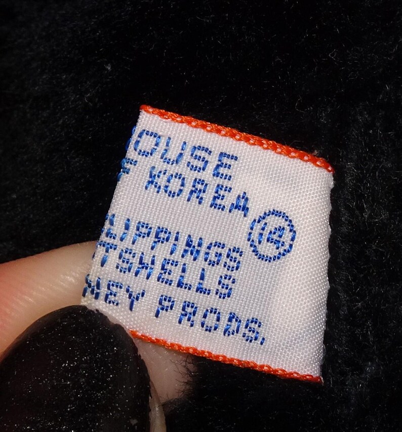 May include: A white label with blue text and an orange border is held against a black background. The label reads "HOUSE OF KOREA" and "LIPPINGS TENELLS KEY PRODS."
