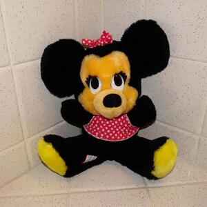 May include: A plush Minnie Mouse doll with black fur, a yellow face and feet, and a red polka dot bow and dress. The doll has large black ears and is sitting with its legs spread.