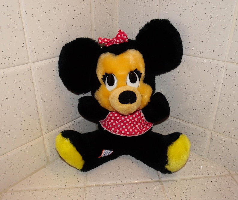 May include: A plush Minnie Mouse doll with black fur, a yellow face and feet, and a red polka dot bow and dress. The doll has large black ears and is sitting with its legs spread.
