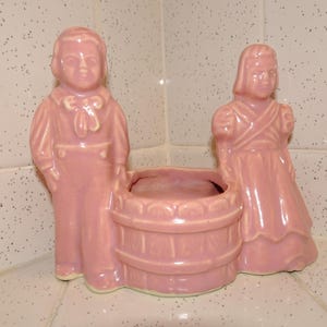 May include: A vintage pink ceramic planter featuring a boy and girl flanking a barrel-shaped container. The figures are dressed in period clothing, and the planter has a glossy finish. The planter is a decorative item.