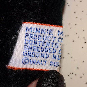 May include: A close-up of a white tag with blue text and an orange border. The tag reads "MINNIE M PRODUCT OF CONTENTS: SHREDDED GROUND NU &copy; WALT DIS". The tag is attached to a black plush toy.