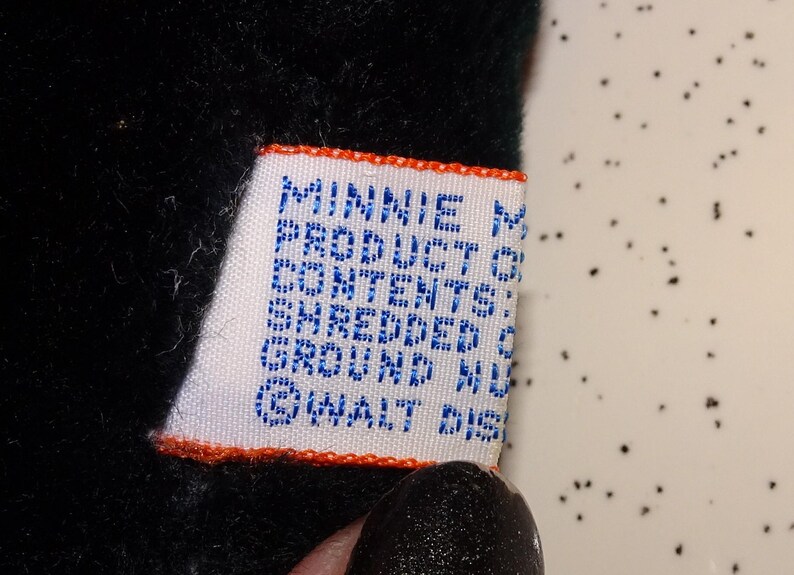May include: A close-up of a white tag with blue text and an orange border. The tag reads "MINNIE M PRODUCT OF CONTENTS: SHREDDED GROUND NU &copy; WALT DIS". The tag is attached to a black plush toy.