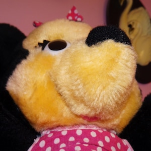 May include: Close-up of a plush toy with yellow fur, black ears, and a black nose. The toy has a pink and white polka dot bow and a pink and white polka dot dress. The background is a solid pink colour.