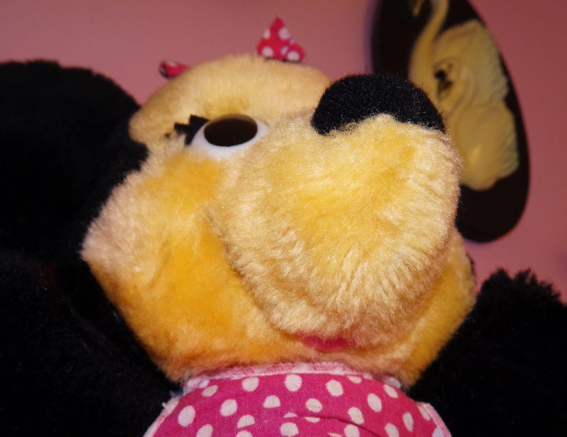 May include: Close-up of a plush toy with yellow fur, black ears, and a black nose. The toy has a pink and white polka dot bow and a pink and white polka dot dress. The background is a solid pink colour.