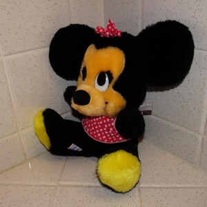 May include: A plush Minnie Mouse doll, with black fur, a yellow face, and large black ears. She wears a red polka-dot bow and matching neckerchief. The doll has yellow feet and is seated.