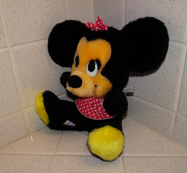 May include: A plush Minnie Mouse doll, with black fur, a yellow face, and large black ears. She wears a red polka-dot bow and matching neckerchief. The doll has yellow feet and is seated.