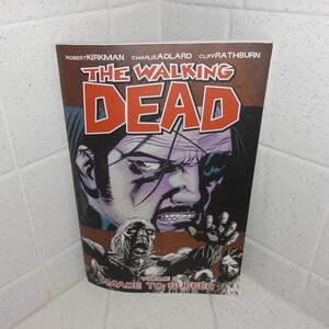 May include: The Walking Dead Volume 8 comic book cover. The cover features a close-up of a man's face with a zombie in the foreground. The title is in large orange letters. The volume is titled "Made to Suffer."