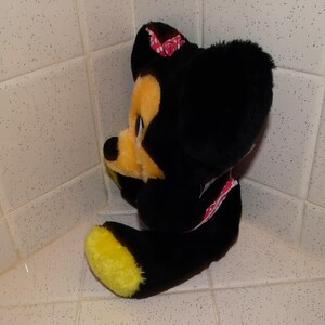 May include: A soft toy mouse with a black body, yellow face and feet, and a pink bow with white spots. The mouse is in a seated position, with its legs stretched out and arms crossed.