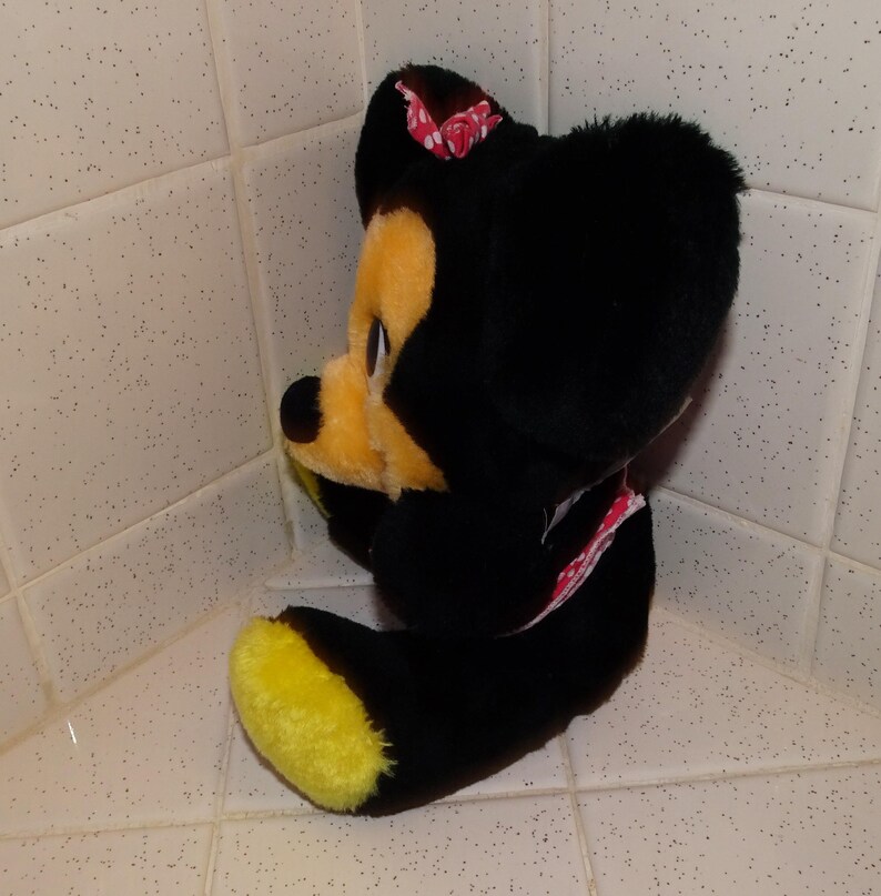 May include: A soft toy mouse with a black body, yellow face and feet, and a pink bow with white spots. The mouse is in a seated position, with its legs stretched out and arms crossed.