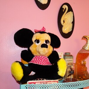 May include: A vintage Minnie Mouse soft toy with black ears, a yellow face, and a red polka dot bow and dress. The toy is on a shelf with other vintage items, including a box of soap and decorative swan plaques.