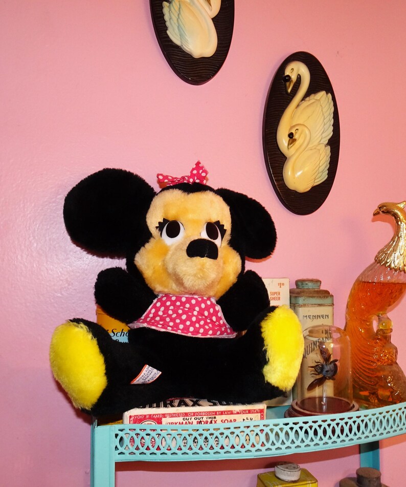 May include: A vintage Minnie Mouse soft toy with black ears, a yellow face, and a red polka dot bow and dress. The toy is on a shelf with other vintage items, including a box of soap and decorative swan plaques.