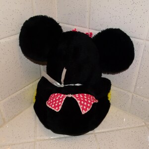 May include: A black plush mouse head toy with large, round ears. It has a red bow with white polka dots and a smaller pink bow on top. The toy is placed in a corner, against a tiled surface.