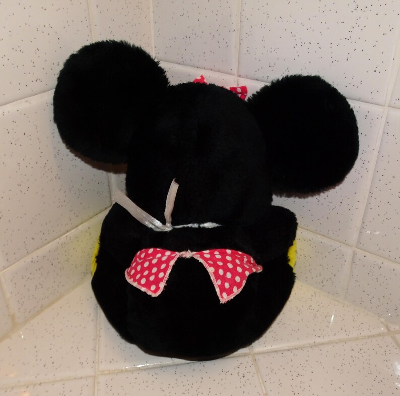 May include: A black plush mouse head toy with large, round ears. It has a red bow with white polka dots and a smaller pink bow on top. The toy is placed in a corner, against a tiled surface.
