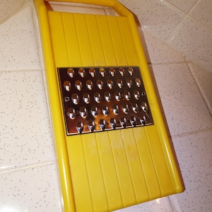Peut inclure: Une râpe en plastique jaune avec une surface de râpage en acier inoxydable. La râpe a une forme rectangulaire avec une poignée en haut. La surface de râpage est en acier inoxydable avec de nombreux petits trous.