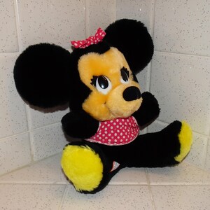 May include: A plush Minnie Mouse doll with black fur, a yellow face and feet, and a red polka-dot bow and dress. The doll has large black ears and big eyes. The doll is sitting in a corner.