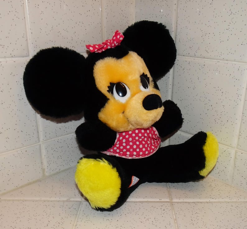 May include: A plush Minnie Mouse doll with black fur, a yellow face and feet, and a red polka-dot bow and dress. The doll has large black ears and big eyes. The doll is sitting in a corner.