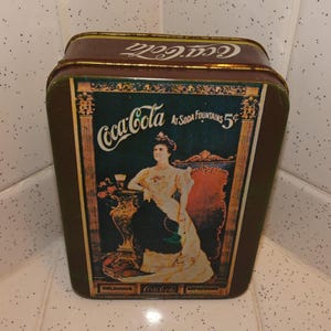May include: Vintage Coca-Cola tin with a brown exterior and a decorative image of a woman in a long dress. The tin features the Coca-Cola logo and the text "At Soda Fountains 5¢".