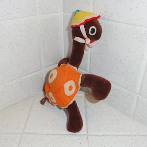 Op de afbeelding: Een vintage pluche schildpad speelgoed. Het heeft een bruin lichaam, een oranje schild met witte cirkels en een gele hoed met rode rand. De schildpad heeft een lange nek en draagt een roze lint. Het speelgoed staat.