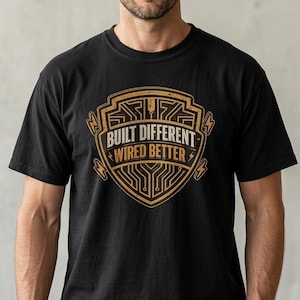 May include: Black t-shirt with a gold and white graphic. The graphic is a shield shape with the words "BUILT DIFFERENT WIRED BETTER" in a stylized font. The design includes lightning bolts and circuit board elements.