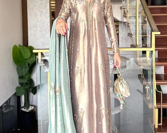Pode incluir: Um vestido túnica comprido até ao chão em tom taupe metálico, com mangas compridas e bordados dourados intrincados no decote, mangas e bainha. A peça é combinada com um xaile dupatta verde menta claro e uma pequena mala de mão dourada a condizer.