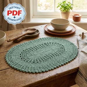 May include: A sage green, oval-shaped crochet placemat with a scalloped edge sits on a wooden table. The placemat has a detailed openwork design in the center. The image also shows wooden spoons, plates, and a small plant in a pot. The text "INSTANT PDF DOWNLOAD" is in a blue and white badge.