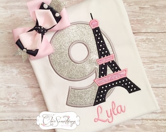 May include: A white t-shirt with a silver glitter number 9 and a black and pink Eiffel Tower design. The name "Lyla" is embroidered below the design. A pink and black polka dot bow is on the shirt.